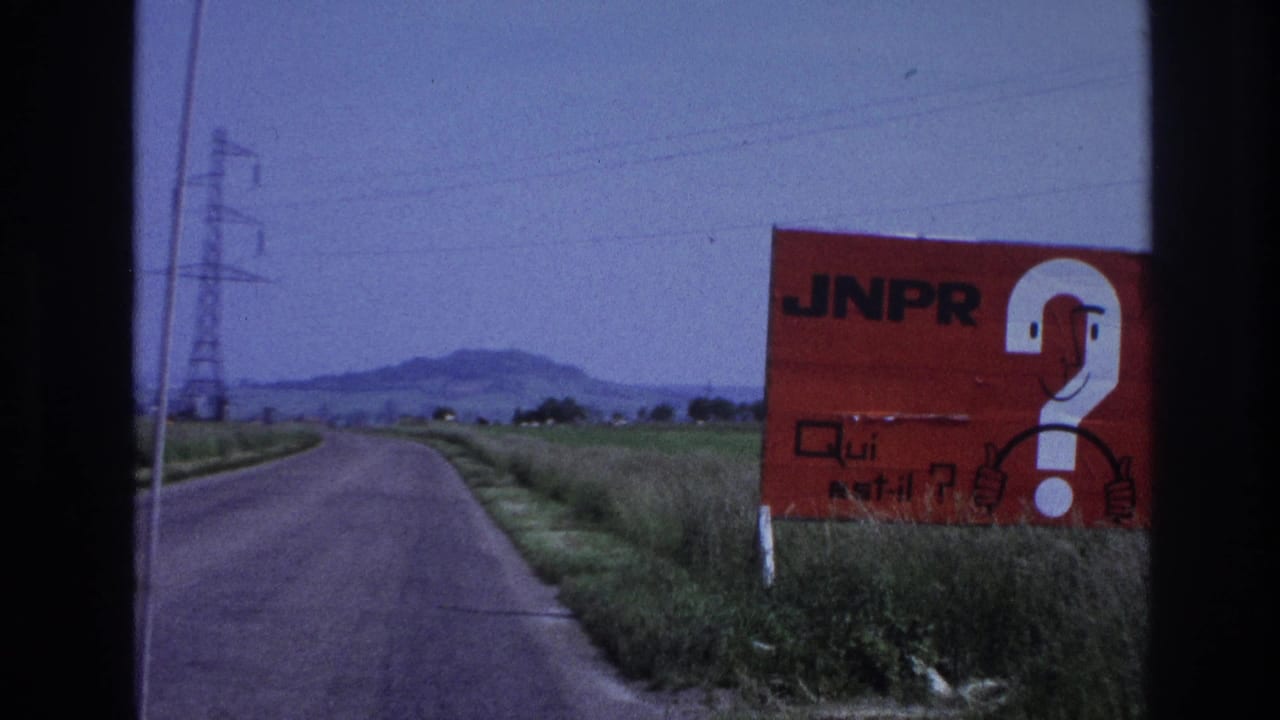 A large red billboard bearing 'JNPR' and a white question mark stands beside a rural French road, circa 1967, photographed through a car windshield on 8mm film.