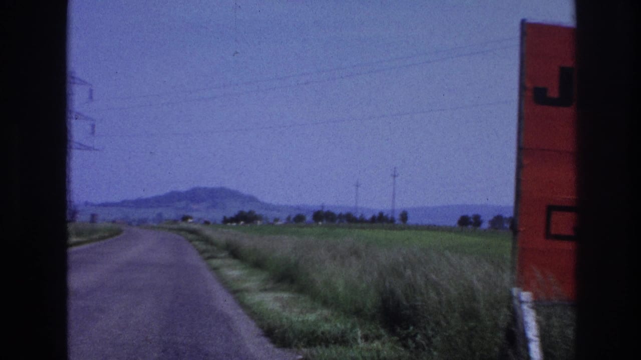 Right edge of a red billboard visible as a vehicle passes on a rural road, open fields, power pylons, and low hills in background.