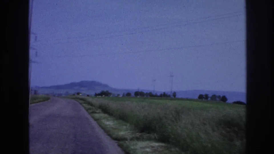 Open two-lane road through rural France after passing the billboard; green verges, power-transmission pylons, and low hills under a hazy sky, circa 1967.