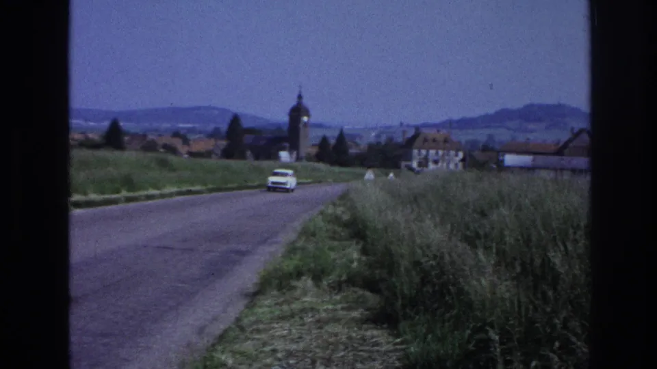 A white period car drives toward a village with a church steeple; green fields and hills in background, rural France, circa 1967, captured on 8mm film.