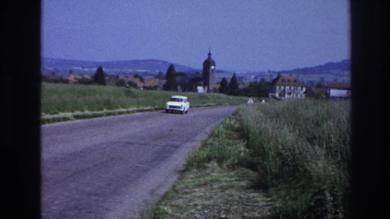 White 1960s station wagon recedes along a rural French road; church tower and rolling hills visible in background, circa 1967, 8mm film.