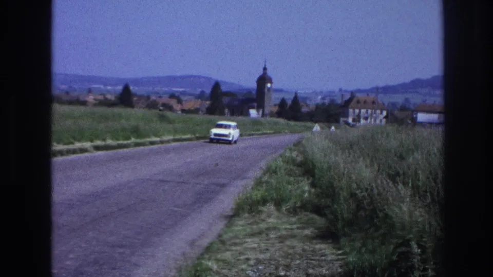 White 1960s station wagon recedes along a rural French road; church tower and rolling hills visible in background, circa 1967, 8mm film.