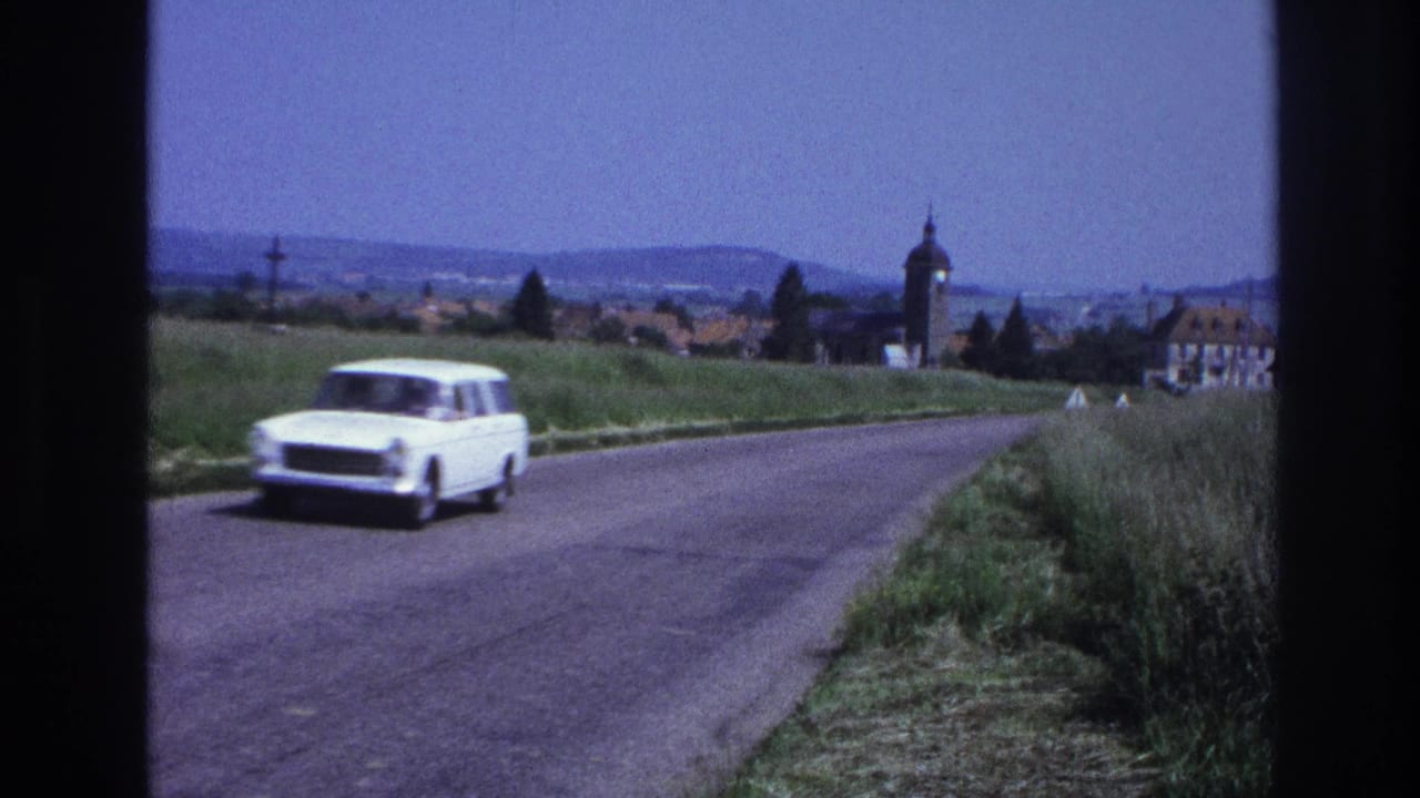 White 1960s European estate car passes close to camera on a rural French road; village, church tower, and rolling hills visible beyond, circa 1967, 8mm film.
