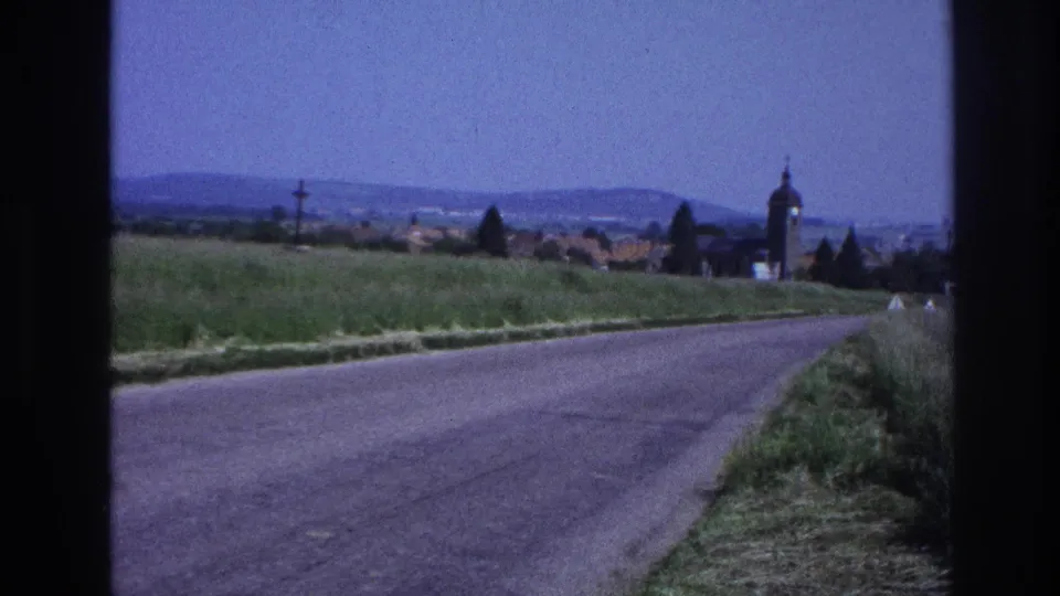 Rural French road, empty after the car has passed; village with square church tower and terracotta-roofed buildings set against rolling hills, 8mm film, circa 1967.