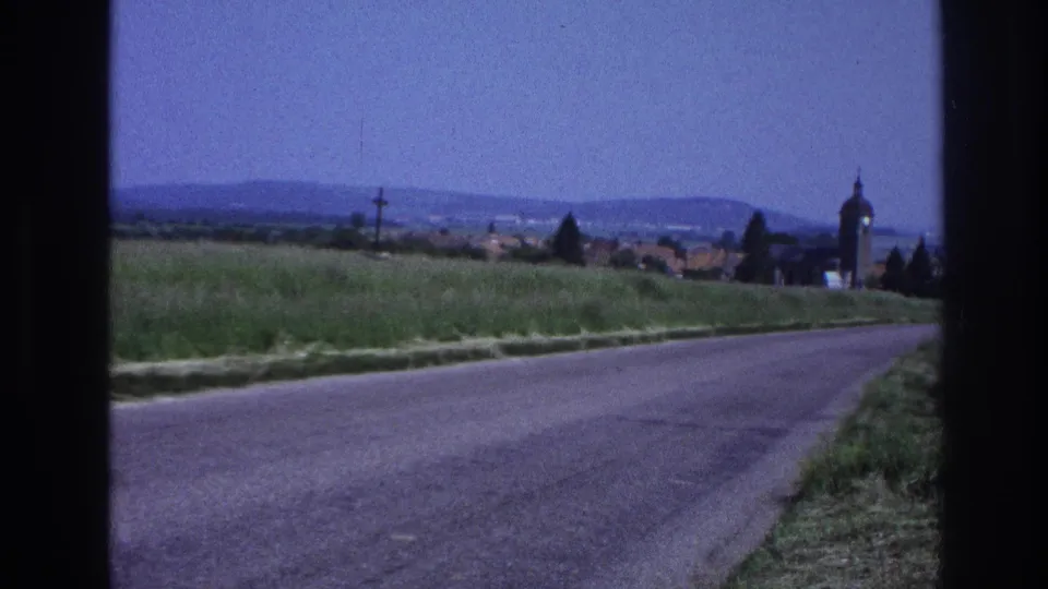 Static roadside shot of an empty rural French road; village church tower and hills remain as film color shifts cooler, 8mm, circa 1967.