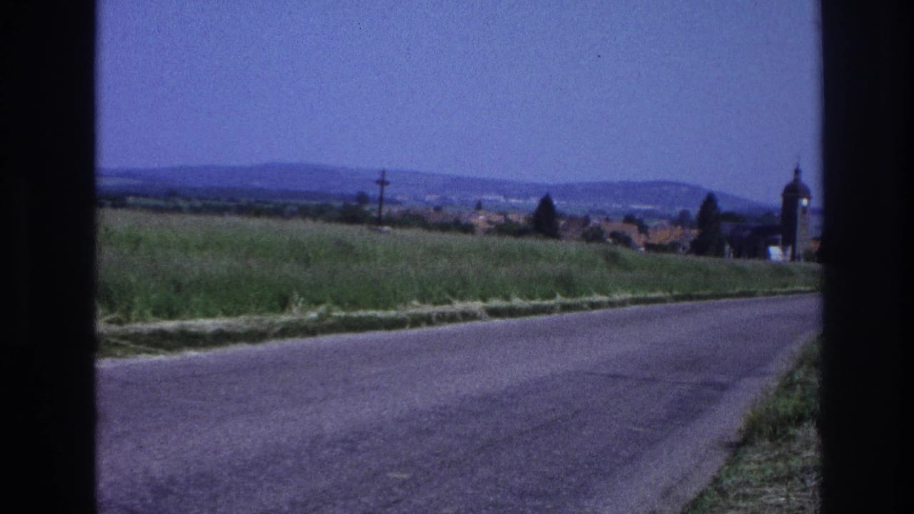8mm home movie from France, c.1967: a country road leads toward a small village with a rounded church tower and orange-tiled rooftops under a blue sky.