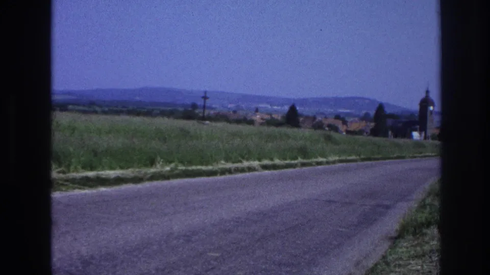 8mm home movie from France, c.1967: a country road leads toward a small village with a rounded church tower and orange-tiled rooftops under a blue sky.