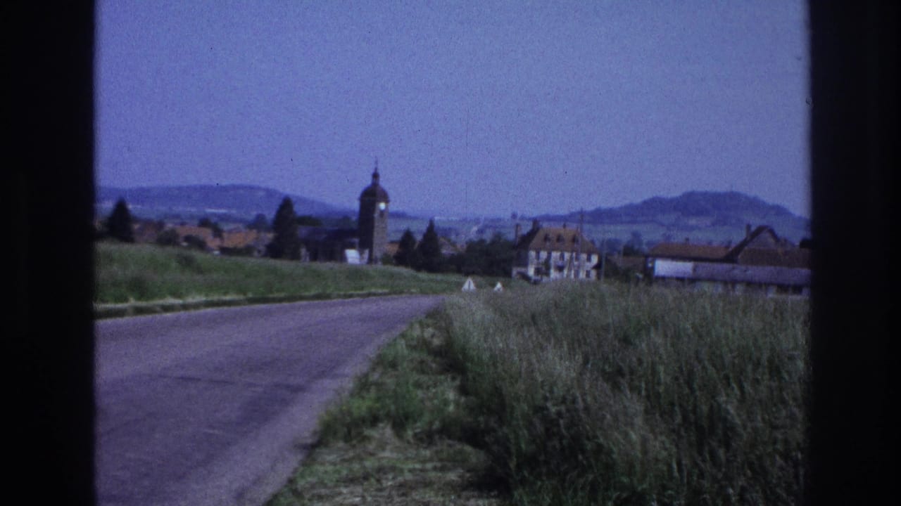 Moving-vehicle POV along a rural road as a French village with a rounded church tower and red-roofed buildings comes into view on the right.
