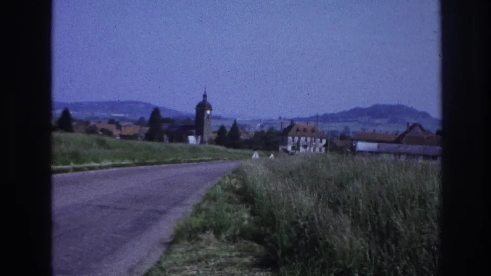 8mm footage from France, c.1967: driving shot as a village with a rounded church tower and traditional orange-tiled buildings appears ahead on the right.