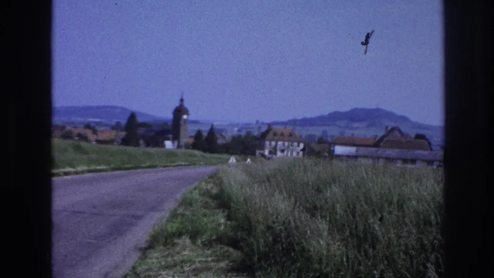 8mm home movie, France c.1967: village with a round-topped church tower, traditional rooftops, green verge, distant hills, and a bird in flight caught mid-frame.