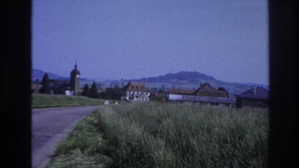 8mm film, France c.1967: the village church tower with a distinctive rounded top dominates the frame, orange-tiled roofs and rolling hills extending behind.