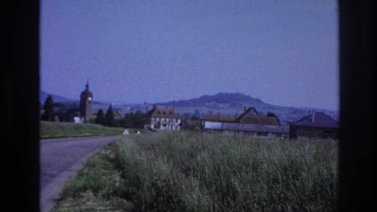 8mm home-movie frame circa 1967, France — roadside grasses and gravel road edge, village rooftops, a domed church tower on the left, and distant hills under a pale blue sky.