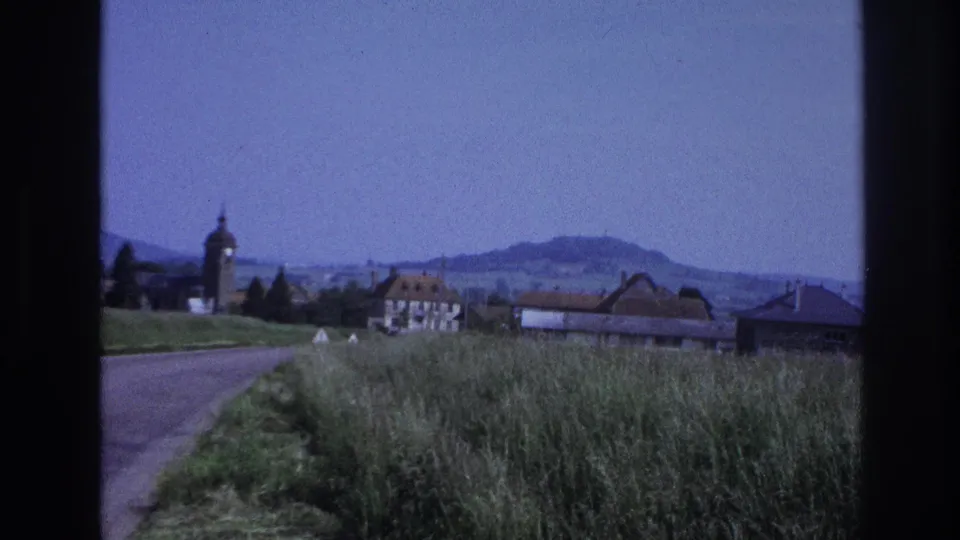 8mm home-movie frame circa 1967, France — roadside grasses and gravel road edge, village rooftops, a domed church tower on the left, and distant hills under a pale blue sky.