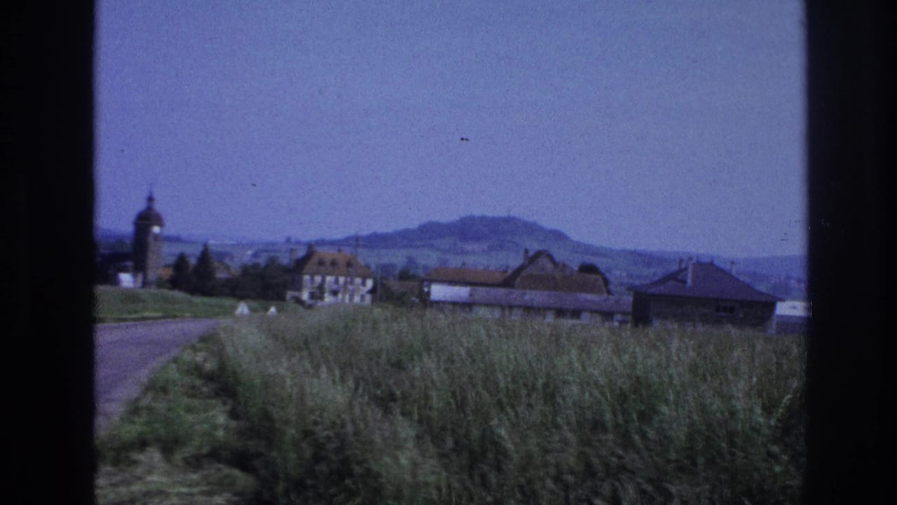 Moving-vehicle shot of a village — tall grasses in foreground, terracotta-roofed houses, a domed church tower on the left, and hazy hills in the distance.