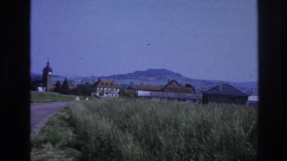 8mm home-movie frame approximately 1967, France — vehicle-shot view of village farmhouses, a rounded church tower still visible left of frame, and rolling hills under blue sky.