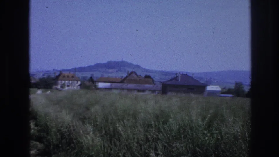 8mm home-movie frame circa 1967, France — church tower now out of frame as vehicle passes; dense grasses, farm buildings, and rounded hills under a clear blue sky.