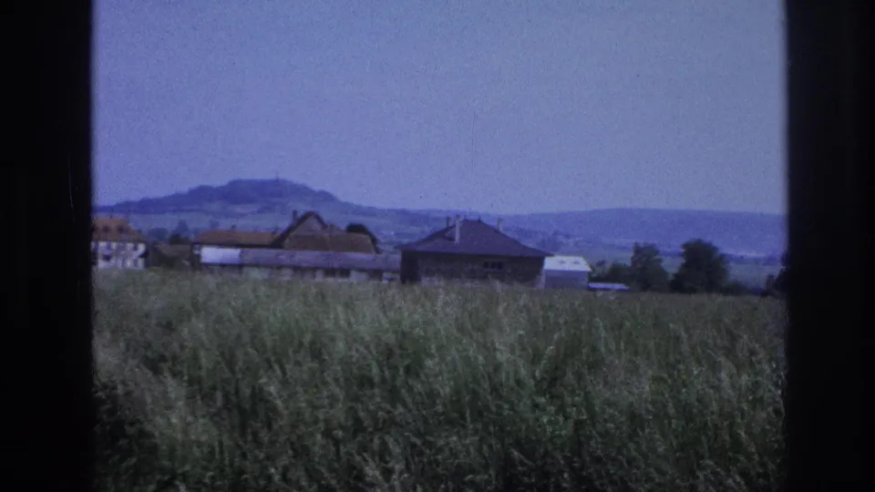 8mm home-movie frame circa 1967, France — final vehicle-shot view; tall grasses, village farm buildings, and distant hills under a clear blue sky with no identifiable landmarks.