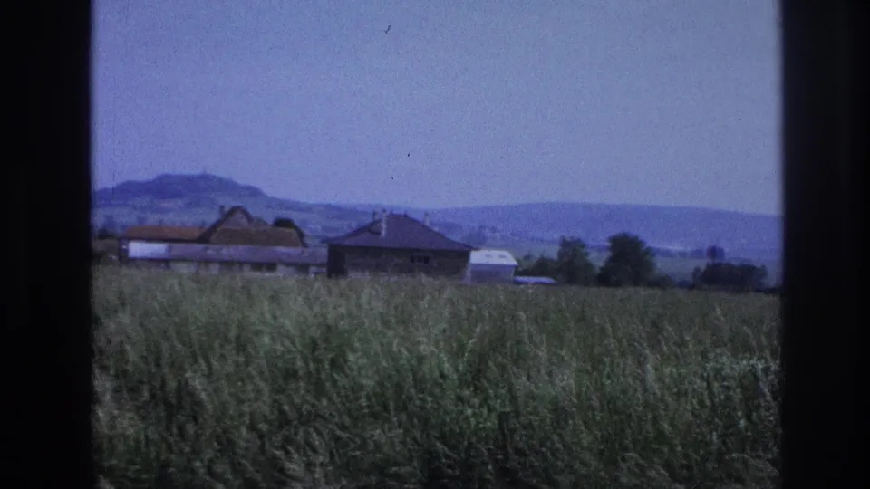 8mm home movie frame showing a dense grass field with a farm barn and rural house in the midground and low hills in the background, France, 1967.
