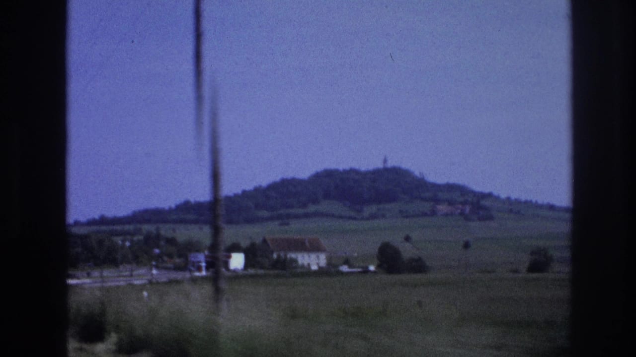 8mm home movie frame of a French rural valley showing a small village, open green pastures, and a prominent wooded hill in the distance, France, 1967.