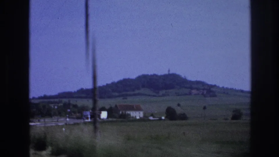 8mm home movie frame of a French rural valley showing a small village, open green pastures, and a prominent wooded hill in the distance, France, 1967.