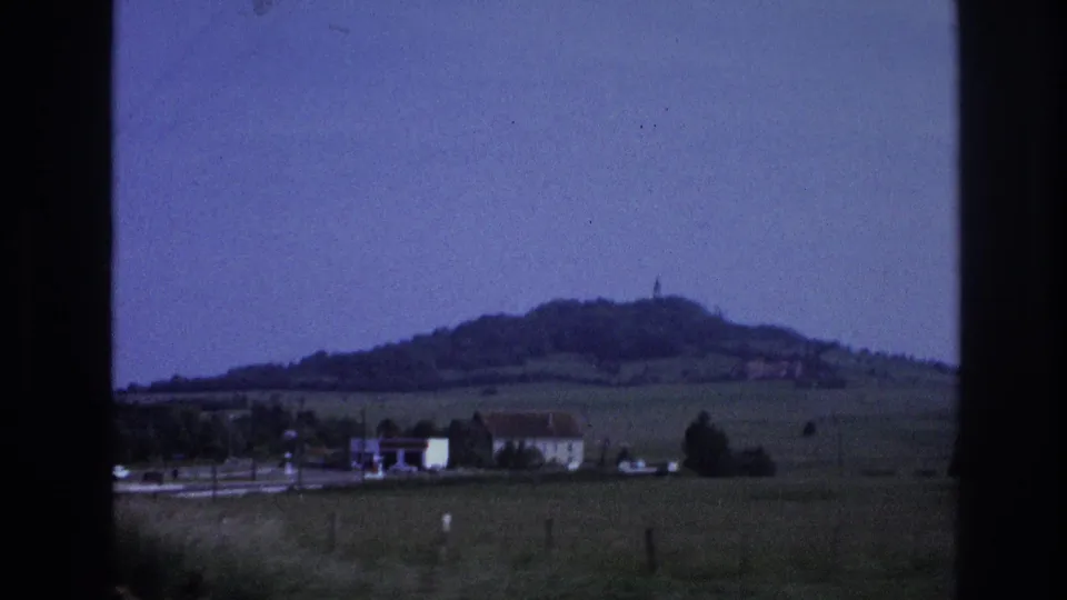 8mm home movie frame from a moving vehicle showing a small French village with red-roofed buildings, open farmland, and a rounded wooded hill in the distance, circa 1967.
