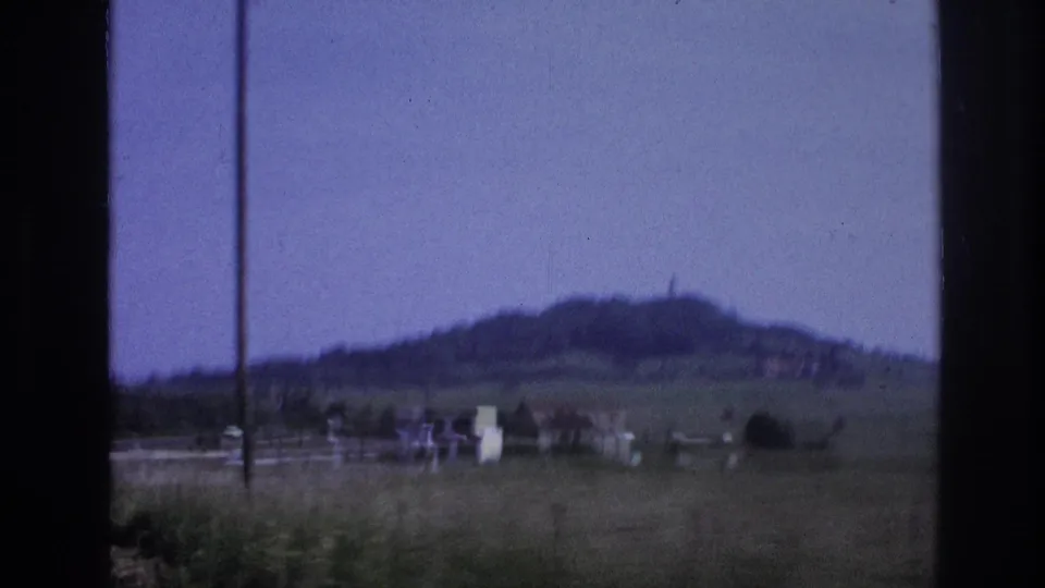 8mm home movie frame of an open French meadow with a distant village cluster and wooded hill, captured from a moving vehicle, France, 1967.