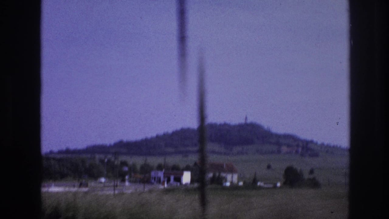 1967 French countryside at passenger-window height: flat green fields, farm buildings, and a rounded hilltop with a small summit structure; 8mm film with pronounced purple dye shift and vertical scratch.