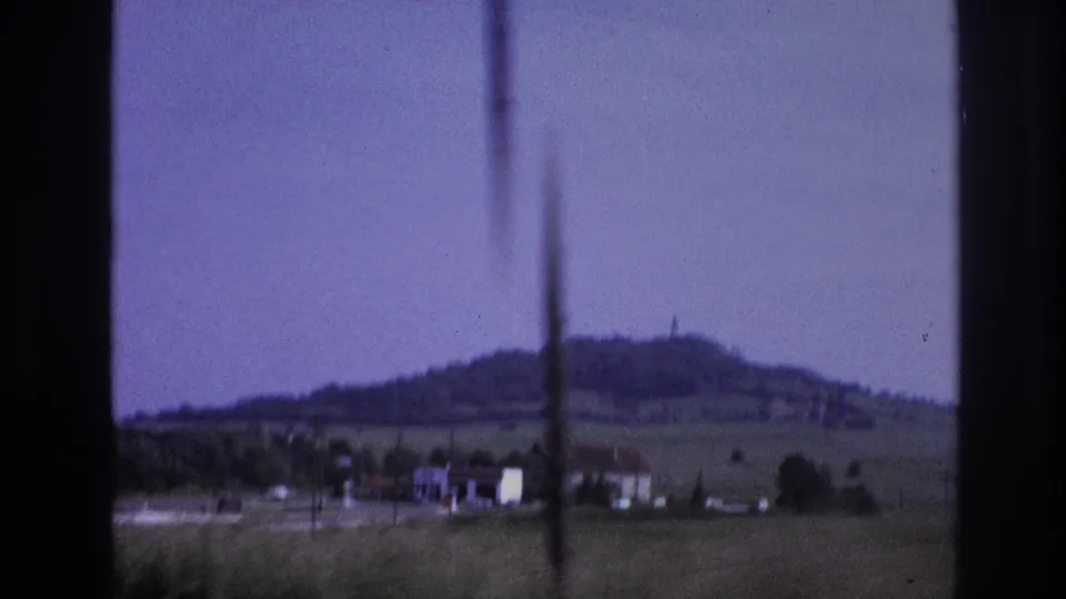 1967 French countryside at passenger-window height: flat green fields, farm buildings, and a rounded hilltop with a small summit structure; 8mm film with pronounced purple dye shift and vertical scratch.