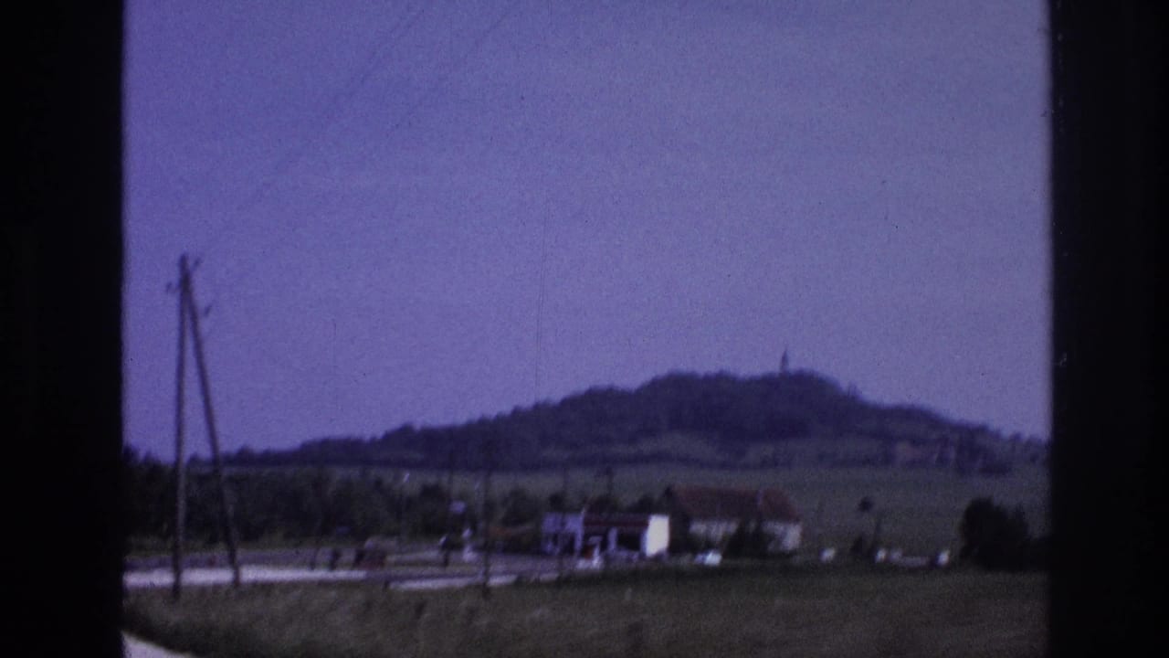Moving vehicle view of French roadside with wooden utility poles in foreground, farm buildings and rounded hill in background, lavender sky.