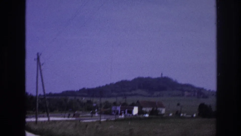 France 1967 roadside at speed: wooden utility poles pass as green farmland and a rounded hill with summit structure recede behind scattered farm buildings; 8mm film with lavender dye shift.