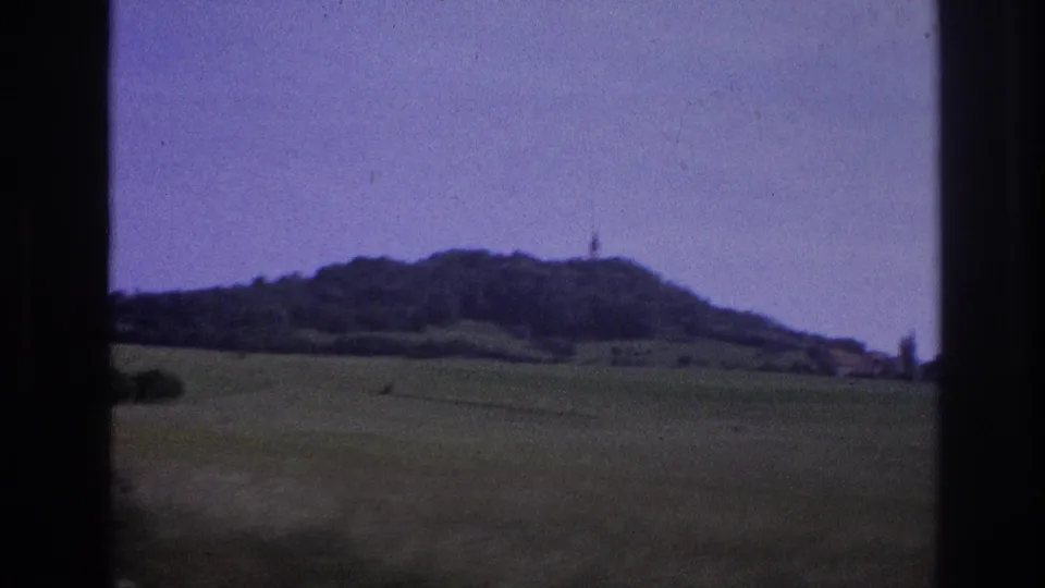 Mid-clip frame, France 1967: wide agricultural fields give way to rolling hills and a distant hilltop bearing a small structure; 8mm film grain and lavender dye cast throughout.