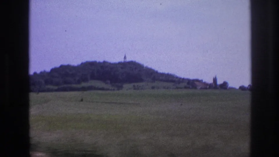 Closing frame, France 1967: a rounded hill with a possible church or tower spire on its summit and buildings clustered at its base; 8mm lavender dye shift, shot from a moving vehicle.