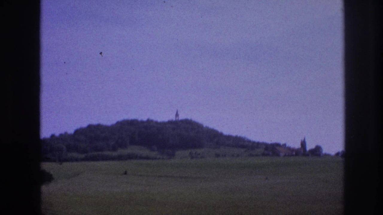 Rolling green hills and wooded hilltop with a spire, framed by a car window on 1960s color film.