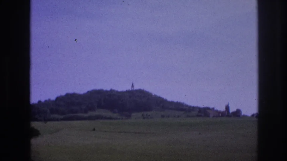 1967 8mm home movie frame: rolling French countryside with a wooded hill topped by a distant spire, shot through a car window with a purple color cast.