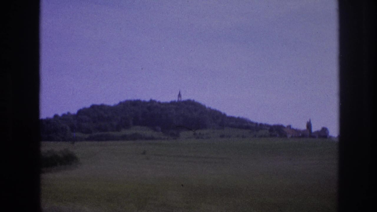 1967 8mm frame: wooded hilltop with a summit tower and a second church spire visible at the right, purple color cast from 1960s film stock, car window at right edge.