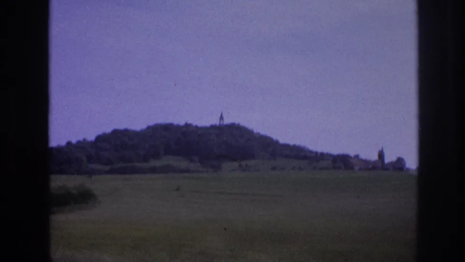 1967 8mm frame: wooded hilltop with a summit tower and a second church spire visible at the right, purple color cast from 1960s film stock, car window at right edge.