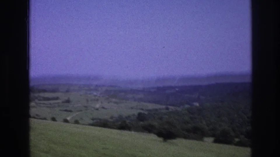 1967 8mm home movie: open valley with undulating green hills receding to a hazy horizon, dark car window and treetop silhouette at lower left, heavy lavender color cast.