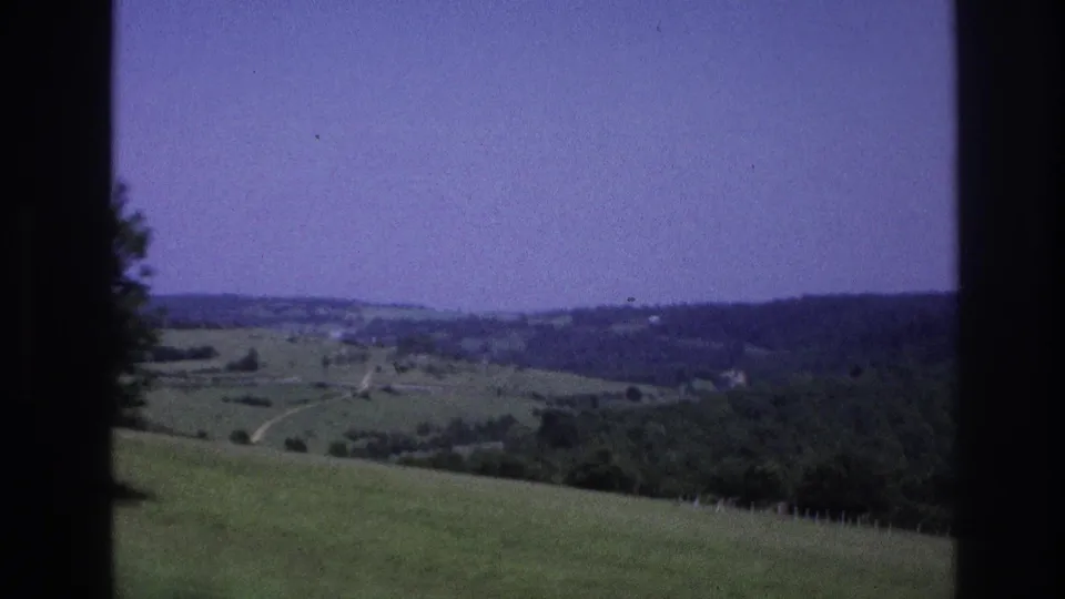 1967 8mm frame: gently rolling countryside with a visible winding road, scattered trees, and small distant settlement, purple film cast, car window pillar at left.