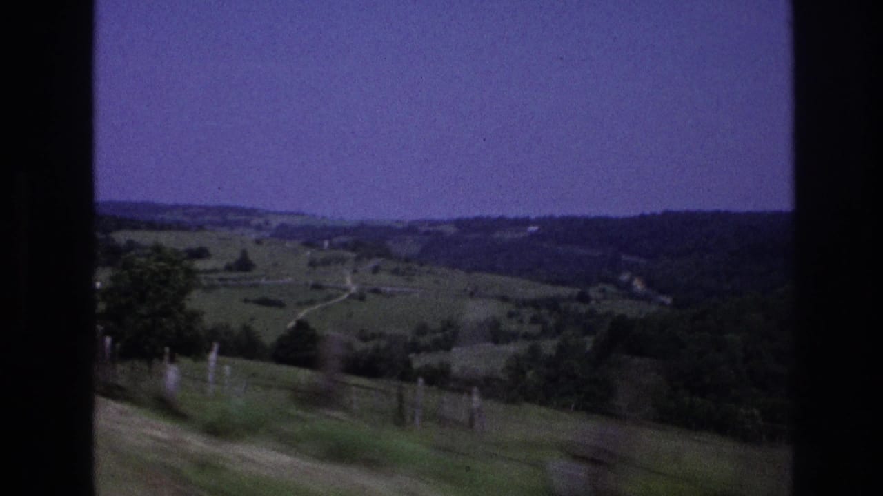 8mm home-movie frame from rural France, 1967, showing rolling green pastures, roadside fence posts, a winding track, and a purple-shifted sky over a tree-lined ridge.
