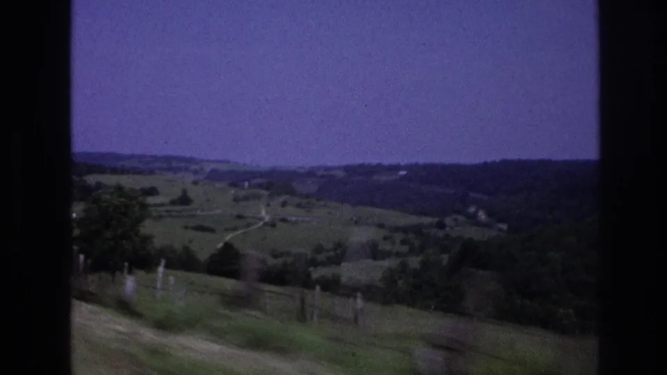 8mm home-movie frame from rural France, 1967, showing rolling green pastures, roadside fence posts, a winding track, and a purple-shifted sky over a tree-lined ridge.