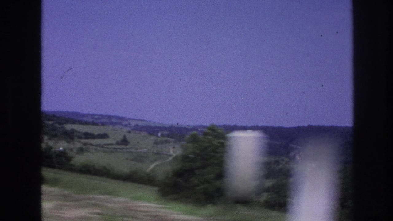 8mm frame from a moving vehicle in rural France circa 1967, showing a green hillside, scattered trees, a blurred white foreground element, and a pale purple sky.