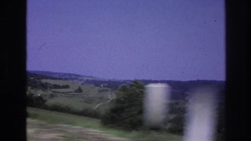 8mm frame from a moving vehicle in rural France circa 1967, showing a green hillside, scattered trees, a blurred white foreground element, and a pale purple sky.