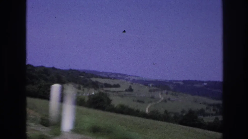 8mm home-movie frame from rural France, 1967, showing a wide pastoral valley, blurred white road marker posts, distant farmland, and a film-dust speck against a purple-shifted sky.