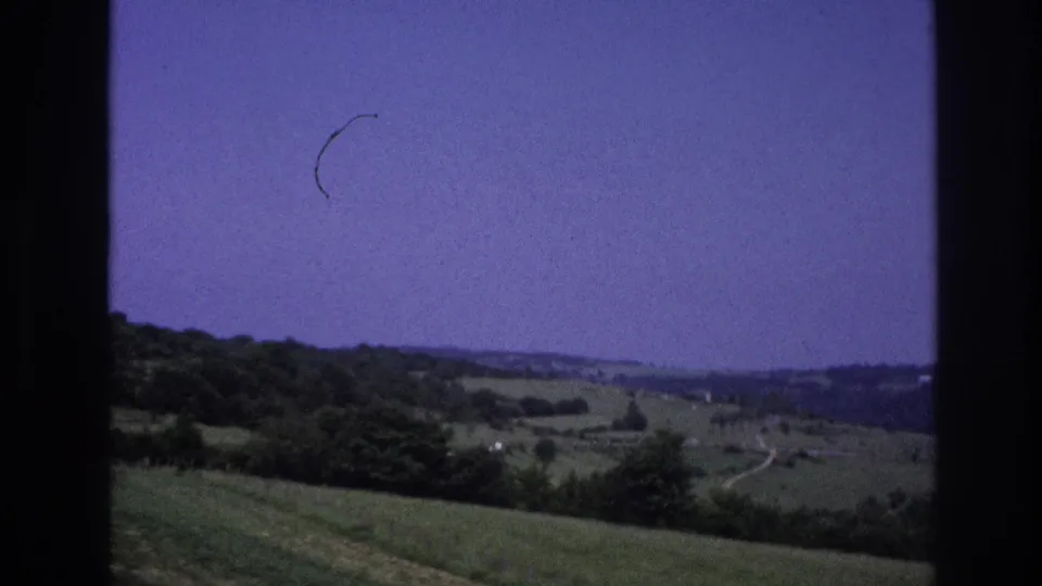 8mm home-movie frame from rural France, 1967, showing pastoral countryside, a curved film-hair scratch in the sky, and winding tracks and scattered trees visible across distant hills.