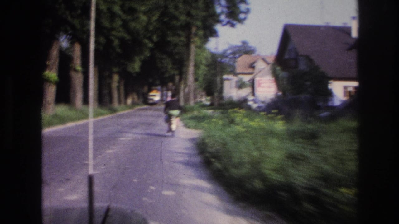 Cyclist on a tree-lined road, white house and yellow vehicle in background; aged 8mm film with blue-purple color shift.