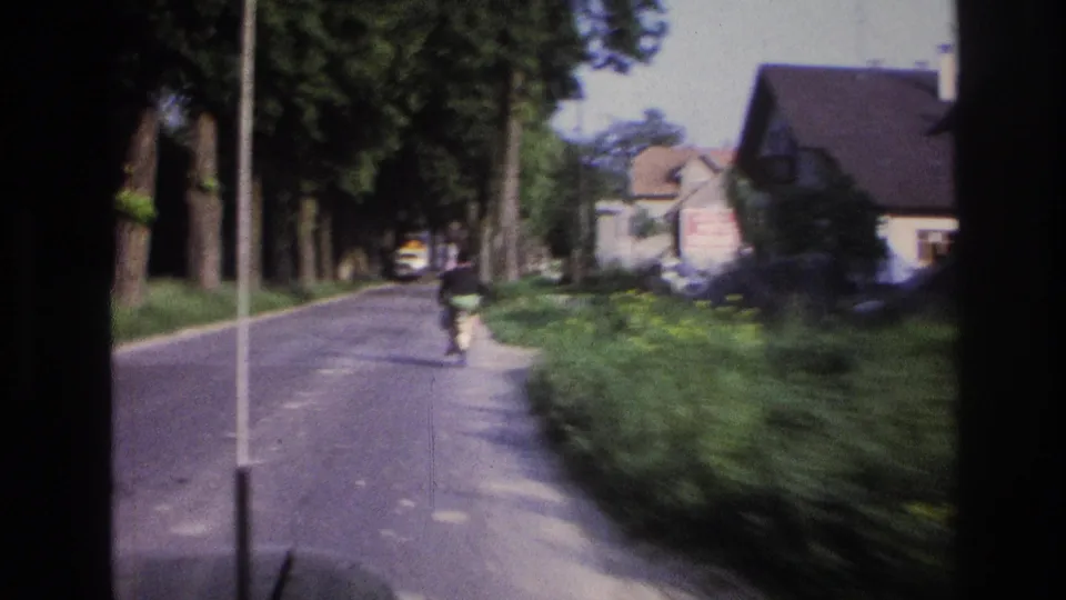 Person cycling away from camera on a tree-lined rural road, white house and yellow vehicle visible in background; appears to be France circa 1967 on aged 8mm film.