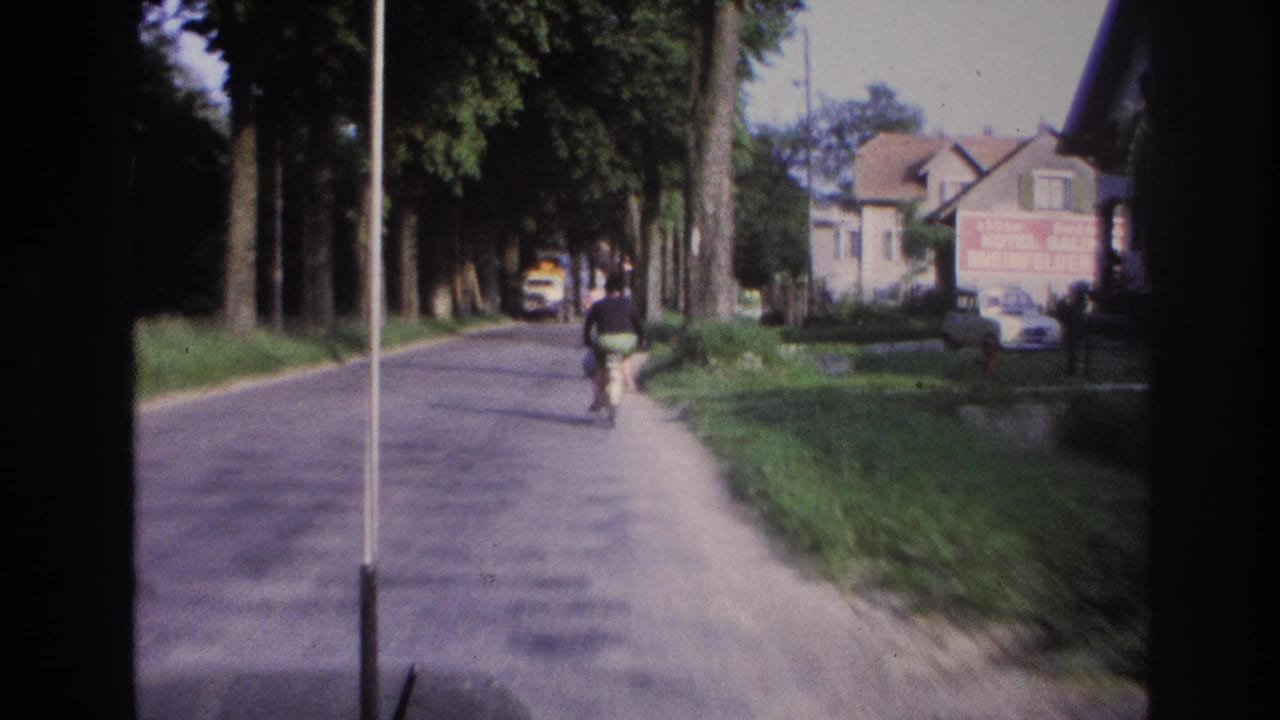 Person on bicycle on a tree-lined village road, house with red commercial signage and yellow vehicle in background; rural France circa 1967 on aged 8mm film.