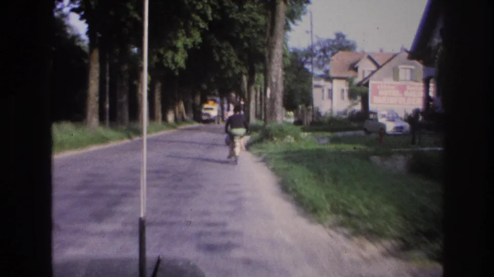 Person on bicycle on a tree-lined village road, house with red commercial signage and yellow vehicle in background; rural France circa 1967 on aged 8mm film.