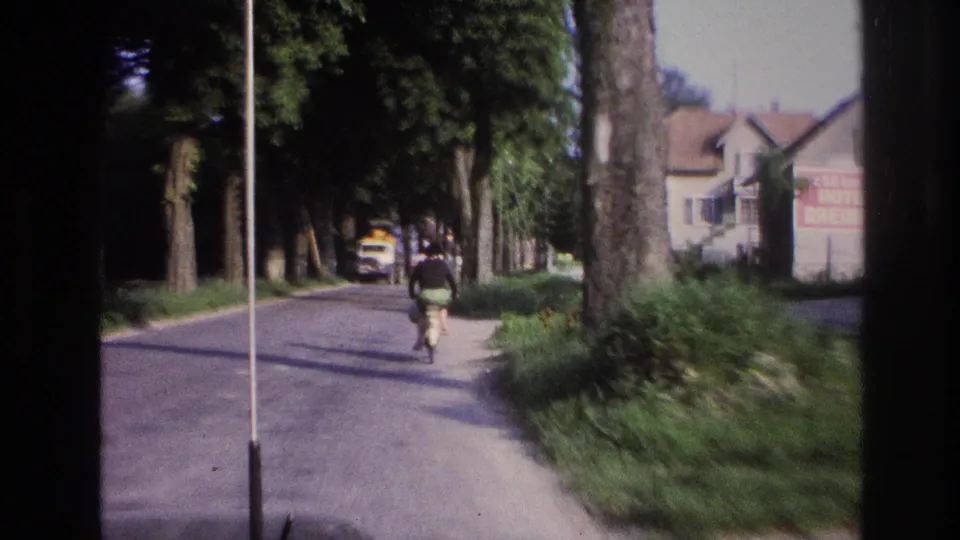 Adult cyclist seen closer from behind, riding a tree-lined village road; yellow truck and red commercial sign visible in background; rural France circa 1967, motion blur from moving vehicle.