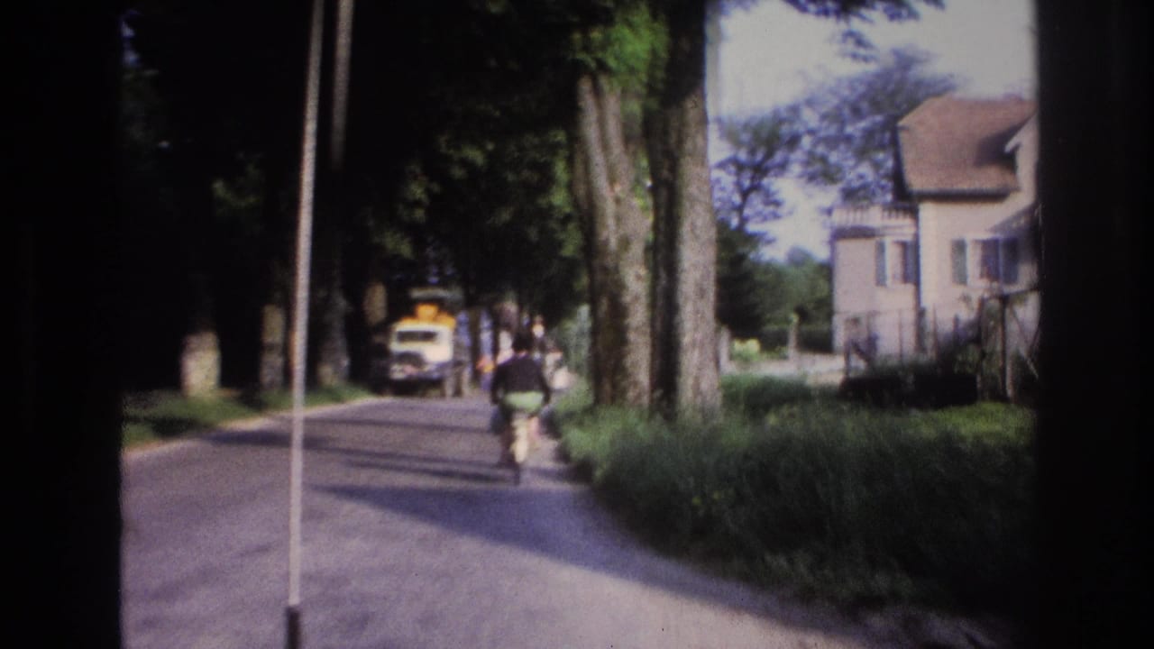 8mm home movie frame, France 1967: male cyclist riding away from camera on a plane-tree-lined road with a yellow van ahead.